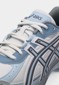 Gros plan sur une chaussure de course Asics bleue et blanche avec des lacets blancs et le logo de la marque sur la languette et les panneaux latéraux.
