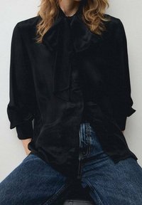 Personne aux cheveux ondulés portant un chemisier noir avec un grand nœud et un jean bleu, assise de manière décontractée les mains dans les poches.