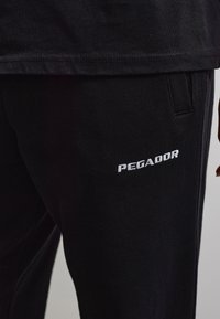 Primo piano di pantaloni neri con tasca e ricamo bianco del logo "PEGADOR" sulla coscia, abbinati all’orlo di una camicia nera.