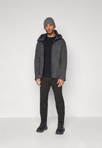Giacca grigia a scacchi con cappuccio, indossata sopra una maglietta nera, abbinata a pantaloni neri e sneakers scure. Berretto grigio di maglia.