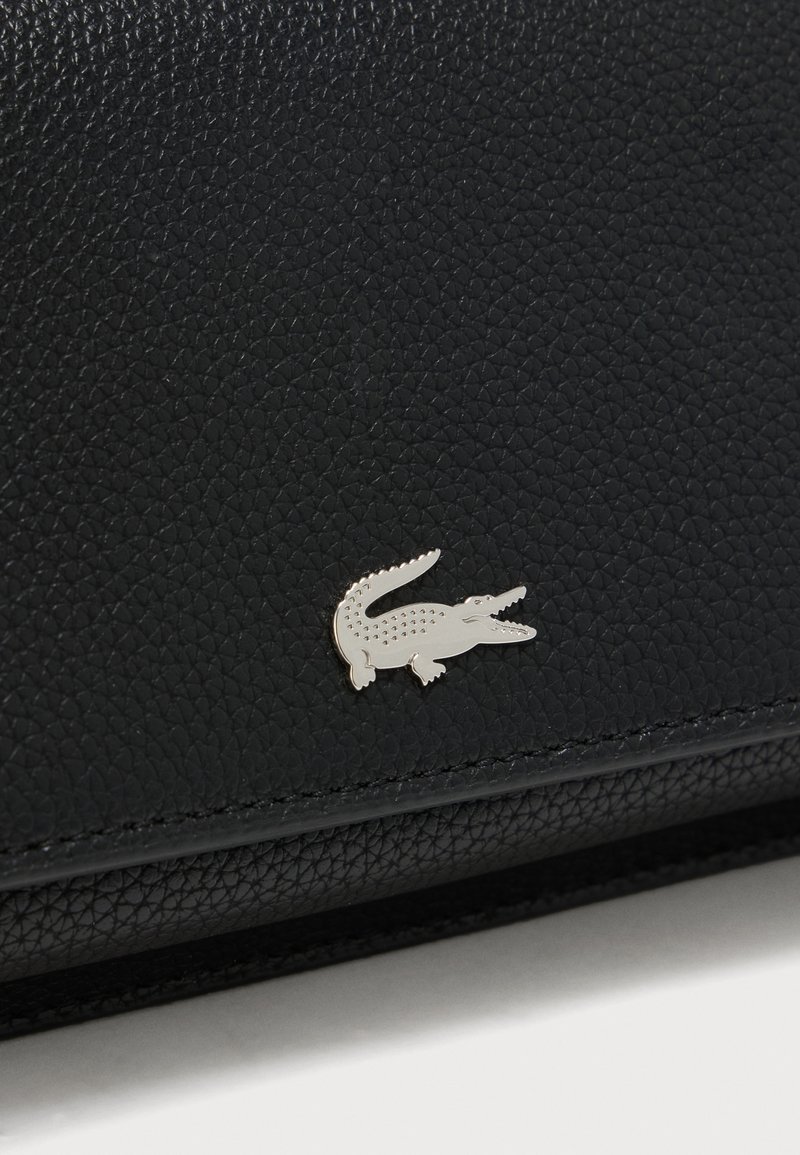 Superficie de cuero negro con textura y un pequeño emblema de logo de cocodrilo plateado adherido cerca del borde inferior.