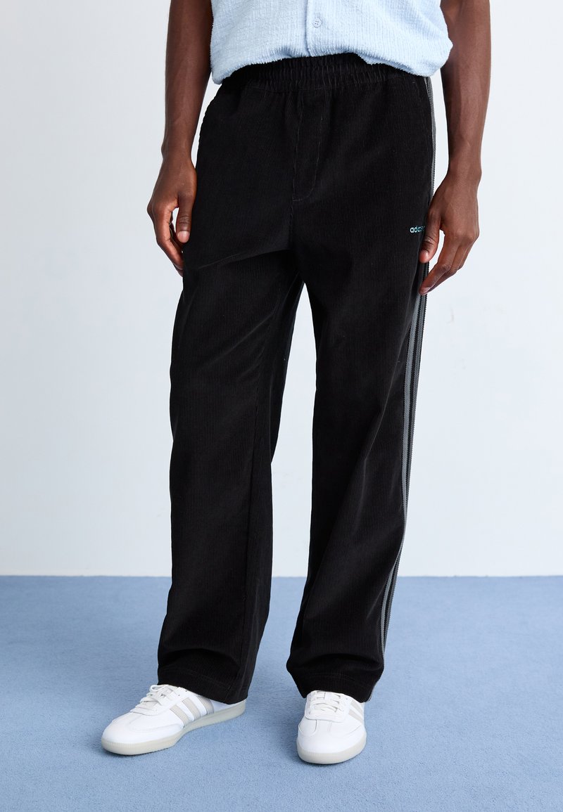 Black corduroy pants with a loose fit, featuring an elastic waistband and contrasting side stripes. Worn with white sneakers.