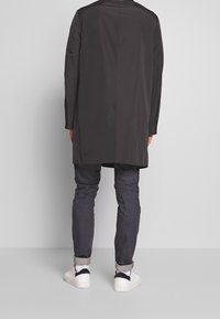 Person iført en sort lang frakke, mørke opsmøgede jeans og hvide sneakers, stående med ryggen til mod en ensfarvet baggrund.