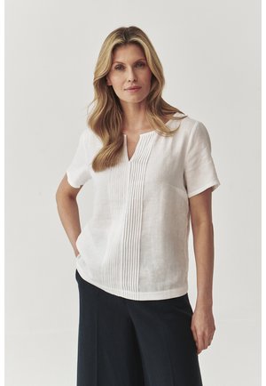 White linen blouse with short sleeves, a keyhole neckline, and vertical pleats down the front. Paired with dark, wide-legged pants.