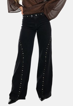 Mujer con vaqueros negros acampanados de talle alto con tachuelas plateadas a lo largo de las costuras frontales y zapatos negros de punta afilada.