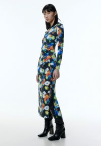 Langärmliges Kleid im lebendigen Blumenmuster mit Blau-, Orange- und Grüntönen. Verfügt über einen seitlichen Schlitz und wird mit schwarzen, knöchelhohen Absatzstiefeln kombiniert.