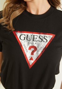 Černé bavlněné tričko s opotřebovaným trojúhelníkovým logem v červené a bílé barvě, na kterém je text "GUESS U.S.A. WASHED JEANS."