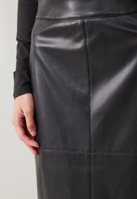 Hand som vilar på en svart läderkjol med synliga sömdetaljer, bärs med en långärmad svart topp mot en enkel bakgrund.