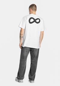 Biała koszulka z dużym czarnym symbolem nieskończoności na plecach, w połączeniu z szarymi dżinsami i jasnokolorowymi sneakery.