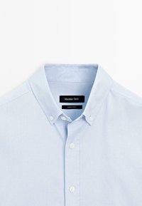 Lichtblauw button-down overhemd met een knoopkraag, drie zichtbare witte knopen en een zwart "Massimo Dutti" label in de kraag.
