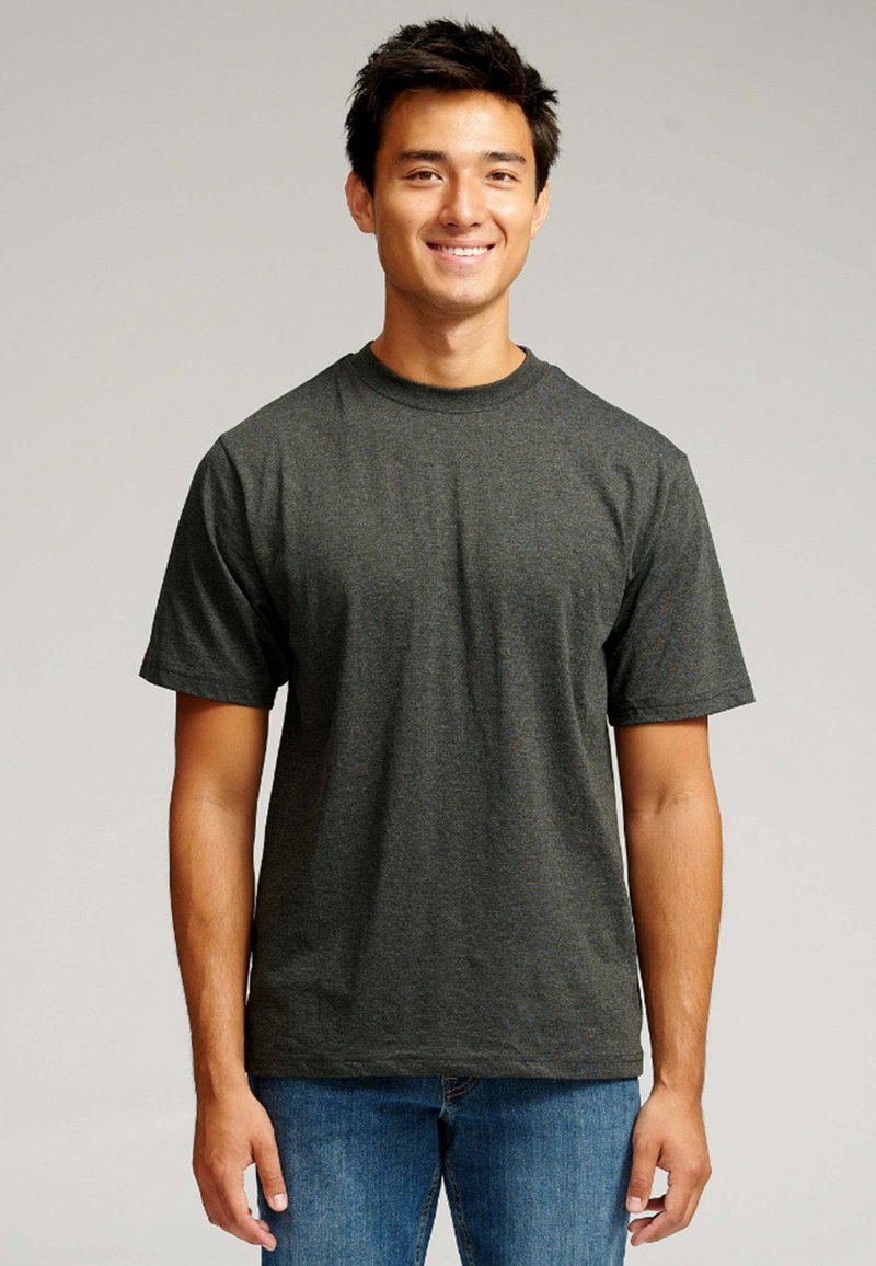 Smilende ung mand med kort mørkt hår, iført en mørkegrå T-shirt og blå jeans, som står foran en enkel lys baggrund.