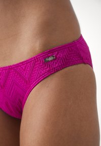 Pink bikini bottom featuring a textured herringbone pattern, with a small metallic logo tag on the waistband, close-up view.