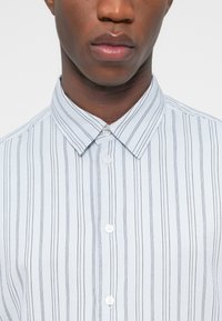 Κοντινό πλάνο ενός ατόμου που φοράει λευκό πουκάμισο με λεπτές κάθετες μαύρες ρίγες και κλασικό γιακά.