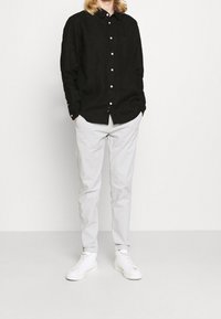 Camicia nera con bottoni e taschino frontale, abbinata a pantaloni grigi chiari a sigaretta e sneakers bianche. La camicia ha un tessuto testurizzato.