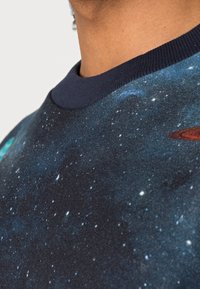 Blauw en zwarte sweater met een ruimteontwerp van sterren en planeten. Geribbelde marineblauwe halslijn, textiel met structuur. Geen zichtbare logo's.