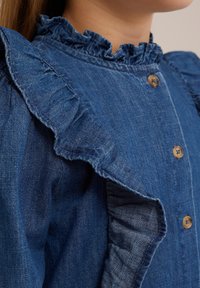 Denimová blůza s řasenými rukávy, tmavě modré barvy, zapínání na knoflíky a jemnou texturou. Obsahuje volánek kolem límce a viditelné prošití.
