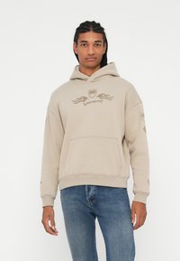 Felpa con cappuccio beige e tasca frontale, logo "VIPER" in marrone chiaro, polsini e orlo a coste. Indossata con jeans in denim blu.