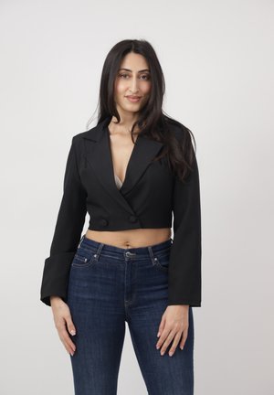 Frau mit langen, dunklen Haaren trägt ein kurzes schwarzes Sakko und blaue Jeans, steht vor einem schlichten hellen Hintergrund und lächelt sanft.