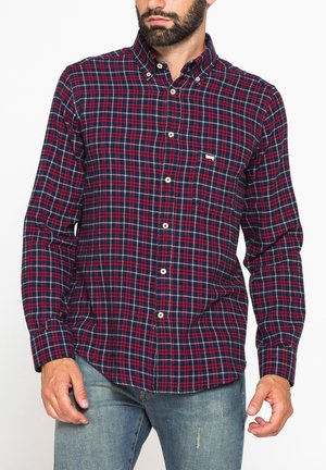 Uomo che indossa una camicia a quadri rossa, blu navy e bianca con taschino sul petto e jeans azzurri usurati, in piedi su sfondo bianco.