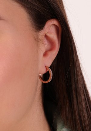 Rozen gouden creolen met een gestructureerd oppervlak en een kleine rij heldere stenen langs de rand. Draagt een model aan het oor.