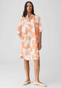 Korte mouw jurk in perzikkleur, met grote witte bloemenprint en een kraagloos ontwerp. Glad materiaal, knie-lengte met zijsplitten.