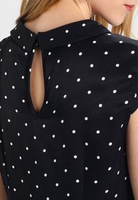 Blusa blu navy con pois bianchi, con colletto e design a apertura a chiave sul retro; realizzata in un tessuto liscio con una texture morbida.