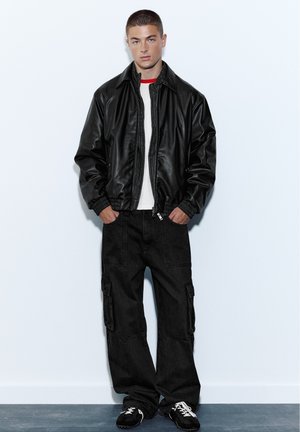 Jeune homme debout, les mains dans les poches, portant une veste en cuir noire, une chemise blanche, un pantalon cargo noir et des baskets noires avec des lacets blancs.