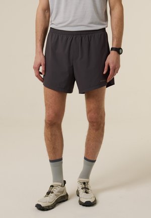 Hombre vistiendo pantalones cortos deportivos negros, calcetines grises, zapatillas de correr beige y un reloj deportivo, de pie contra un fondo liso.