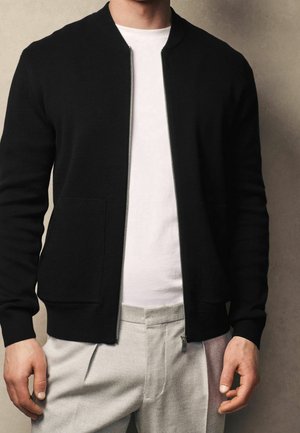 Cárdigan negro de punto con cierre de cremallera, cuello acanalado y dos bolsillos delanteros, llevado sobre una camisa blanca y pantalones gris claro.