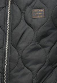 Gestepptede schwarze Jacke mit einem silbernen Reißverschluss, strukturiertem Stoff und einem braunen Etikett mit "JACK & JONES"-Branding auf der Brust.