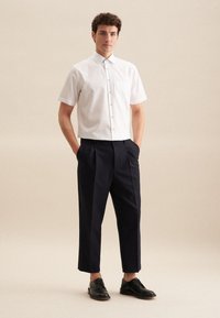 Camicia bianca a maniche corte con bottoni e taschino sul petto, abbinata a pantaloni a pieghe blu navy e scarpe nere con lacci, su uno sfondo neutro.