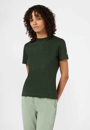 Mujer con cabello rizado, vistiendo una camisa de manga corta verde oscuro y pantalones verde claro, de pie frente a un fondo blanco liso.
