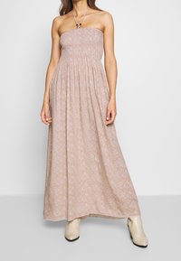 Axel-lös maxiklänning i ljusbeige med blommönster. Rynkad byst, svepande kjol och justerbar nackrem. Buren med beige cowboyboots.