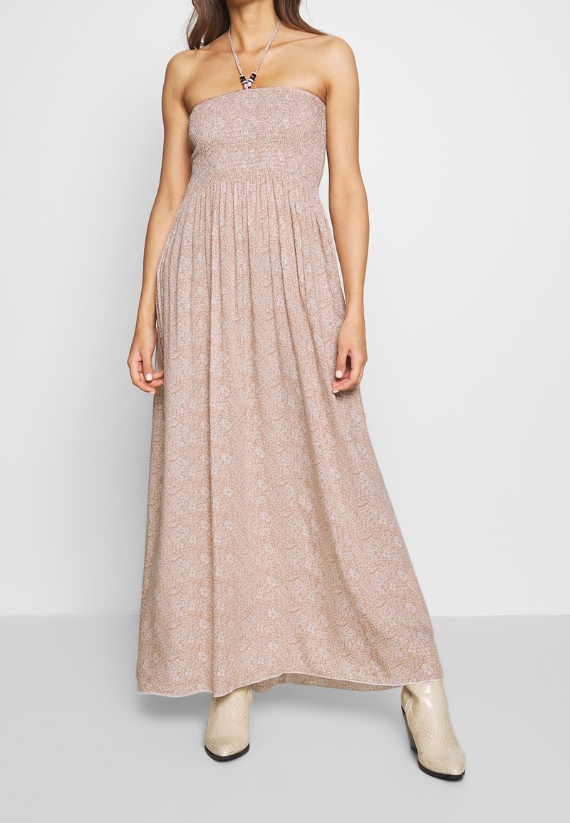 Axel-lös maxiklänning i ljusbeige med blommönster. Rynkad byst, svepande kjol och justerbar nackrem. Buren med beige cowboyboots.