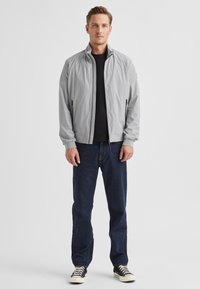 Hellgraue Bomberjacke mit Stehkragen, Frontreißverschluss und Seitentaschen, getragen über einem schwarzen Shirt und dunkelblauen Jeans. Schwarze Turnschuhe.