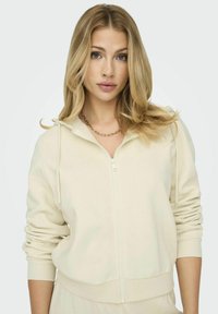 Felpa crema con zip, cappuccio regolabile, polsini e orlo a coste, texture liscia e design minimalista. Indossata con una delicata collana di catena dorata.