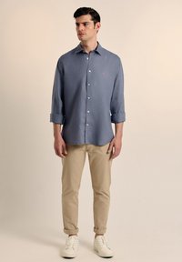 Camisa de botón azul claro con mangas enrolladas, combinada con chinos beige y zapatillas blancas. Presenta un pequeño logo rojo en el bolsillo del pecho.