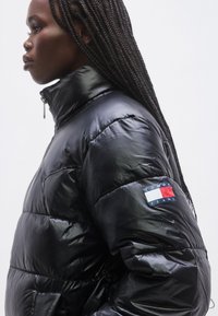 Čierna prešívaná bunda s lesklým vzhľadom, vysokým golierom a zapínaním na zips. Na ľavom rukáve sa nachádza nášivka s logom v červenej, bielej a modrej farbe.