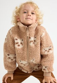 Tan fleece jacket with a soft, textured surface and a zip closure. It features light brown accents and a playful pattern of darker spots.