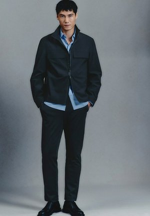 Hombre de pie con las manos en los bolsillos, vestido con chaqueta negra, camisa azul, pantalones negros y zapatos negros, frente a un fondo gris liso.