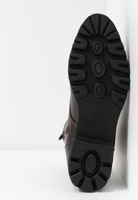 Semelle d'une botte marron foncé avec des crampons profonds, des motifs circulaires et une adhérence texturée, reposant sur un fond blanc.