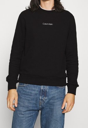 Person, der ein schwarzes Calvin Klein Sweatshirt mit weißem Logo auf der Brust und blauen Jeans trägt, steht mit entspannten Händen an den Seiten.