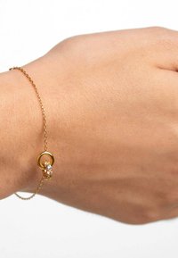 Bracciale in catena d'oro con un ciondolo circolare decorato con piccole pietre preziose trasparenti, indossato su una mano di carnagione chiara. Design semplice, texture delicata.