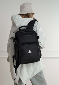 Mochila negra de tela, con múltiples bolsillos con cremallera, un asa superior y correas ajustables para llevar. Detalle de logo triangular.