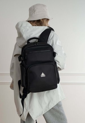 Zaino nero in tessuto, con numerose tasche con zip, una maniglia superiore e spallacci regolabili per il trasporto. Dettaglio logo triangolare.