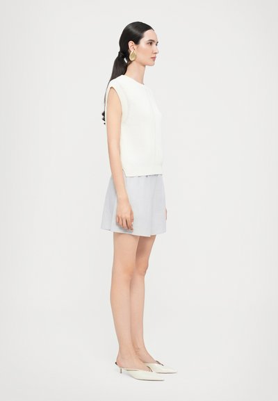 Top smanicato bianco a maglia, abbinato a una gonna grigia chiara. Indossando mule beige, che mostrano una texture liscia e un design minimal.
