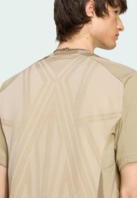 Krátký rukáv béžové košile s texturovaným geometrickým vzorem na zádech, hladkým povrchem látky a kontrastním designem ramene.
