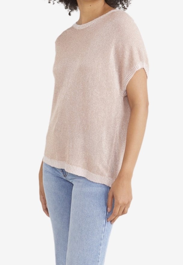 Femme portant un pull ample en maille rose clair à manches courtes, associé à un jean bleu clair, sur un fond blanc.