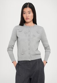Donna che indossa un cardigan grigio lavorato a maglia con decorazioni floreali e bottoni, abbinato a pantaloni scuri a righe sottili, in piedi con le mani nelle tasche.