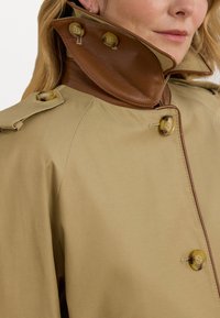 Manteau beige avec un col en cuir marron, deux grands boutons jaunes et une texture lisse. Présente une fermeture à double bouton distincte.
